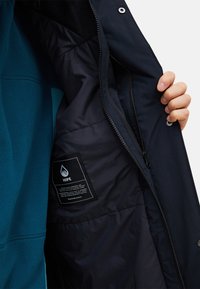 Giacca isolante blu navy con un'esterno liscio e resistente all'acqua. Presenta una tasca con zip e un'etichetta che dettaglia la composizione dei materiali.