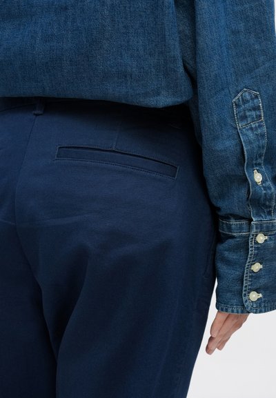 Džinsa krekls ar zilu nokrāsu, ar pogājamām manšetēm un gludu tekstūru, kombinēts ar tumši zilām biksēm ar diskrētu aizmugurējo kabatu.