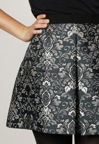 Main sur la taille au-dessus d'une jupe à motifs floraux noirs et argentés avec des plis, associée à un haut noir et des collants.