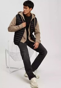 Czarna kamizelka puchowa noszona na beżowej bluzie z kapturem, w połączeniu z ciemnymi dżinsami i beżowymi sneakersami. Postać siedzi na białym metalowym krześle.