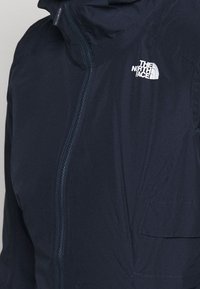 Granatowa wodoodporna kurtka z zamkiem błyskawicznym, kapturem i kieszenią na naszywkę. Posiada białe logo na górnej lewej stronie. Miękka faktura.