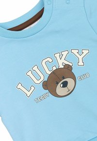 Gaiši zils džemperis ar brūna tedi lāča grafiku un uzrakstu „LUCKY TEDDY CLUB” treknā baltā un melnā drukā.