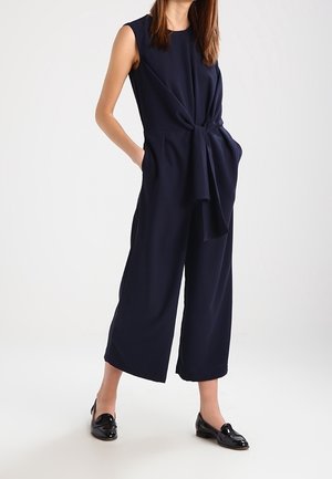 Femme portant une combinaison sans manches bleu marine avec un détail noué à la taille et des mocassins en cuir verni noir, debout les mains dans les poches.