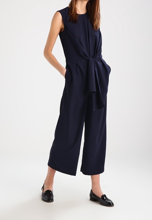 Femme portant une combinaison sans manches bleu marine avec un détail noué à la taille et des mocassins en cuir verni noir, debout les mains dans les poches.