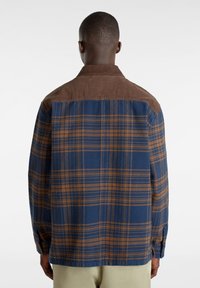 Rutigt bomullsskjorta i blå och brun färgskala, med en solid brunt kordflodertyg vid axlarna och långa ärmar.