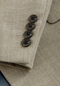 Beżowy, teksturowany rękaw marynarki z czterema ciemnobrązowymi guzikami przyszytymi pionowo w pobliżu mankietu.