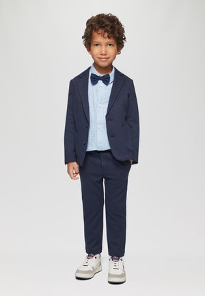 Marineblauwe pak met een op maat gemaakte blazer en bijpassende broek, gecombineerd met een lichtblauw gestreept overhemd en een donkere vlinderdas. Witte sneakers maken de look compleet.
