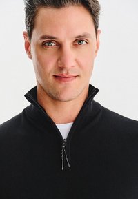 Czarny sweter zapinany na zamek z wysokim kołnierzem, z gładką teksturą i dwoma suwakami, noszony na białej koszuli. Zbliżenie na dekolt.