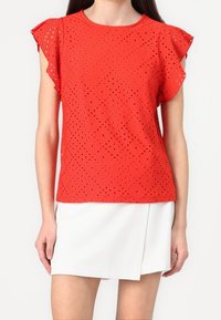 Top corto rosso a maniche corte con design in pizzo a forma di diamante. Collo rotondo, tessuto leggero, delicato dettaglio di ruches alle maniche.