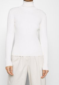 Maglione bianco a collo alto con maniche lunghe, lunghezza fino a metà coscia, realizzato in morbido materiale knit, con un design aderente e superficie testurizzata.