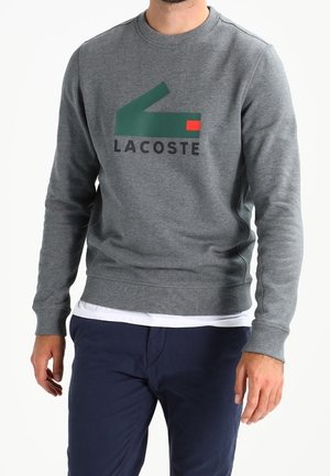 Mężczyzna w szarej bluzie Lacoste z zielonym i czerwonym logo, na białej koszuli i granatowych spodniach.