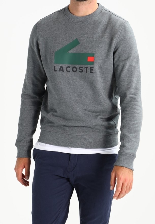 Homme portant un sweat-shirt gris Lacoste avec un logo vert et rouge, superposé à une chemise blanche et un pantalon bleu marine.