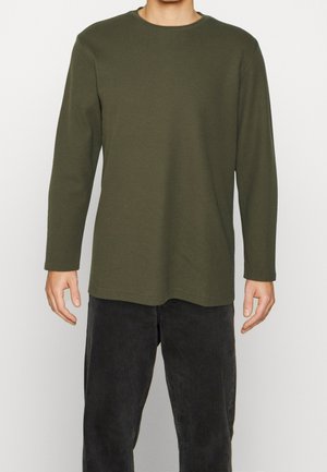 Mann trägt ein schlichtes olivgrünes Langarmshirt und eine schwarze Hose und steht vor einem hellen Hintergrund.