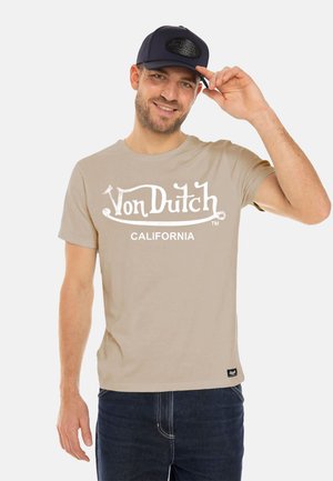 T-shirt en coton beige avec le logo blanc "Von Dutch California", associé à un jean bleu foncé et une casquette marine arborant un patch logo.