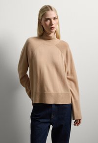 Beige Rollkragenpullover mit gerippten Akzenten, verkürzt geschnitten und mit langen Ärmeln, kombiniert mit dunklen Jeans. Geschmeidige Textur und lockere Passform.