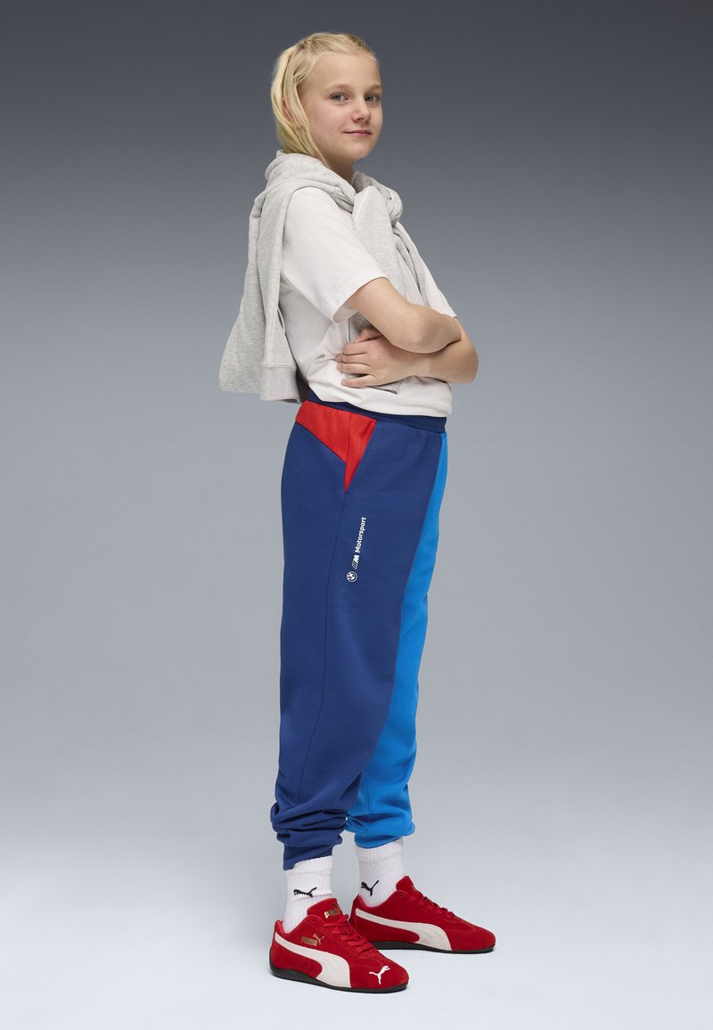 Blaue und rote Color-Blocked-Sweatpants mit einem elastischen Bund, seitlichen Taschen und weißem Logo-Detail, kombiniert mit einem grauen Sweatshirt und roten Sneakers.