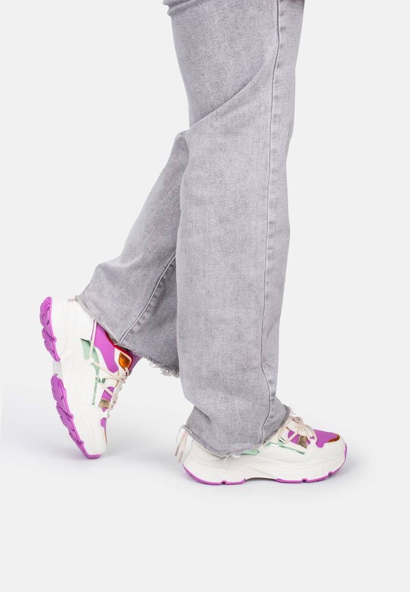 White and purple sneakers with a chunky sole, featuring multicolored accents and decorative laces, worn with gray jeans.