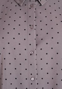 Lilla skjorte med knapper og sorte prikker samt klassisk krave, vist i tæt på detaljer.