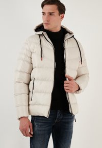 Jachetă puf albă cu glugă, având cusături orizontale, șnururi negre și buzunare laterale cu fermoar. Material texturat și un ușor luciu.