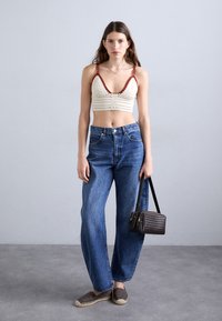 Bralette au crochet crème avec des accents rouges, associé à un jean en denim bleu ample. Le mannequin porte un petit sac à main en osier marron et est stylé avec des chaussures à enfiler.
