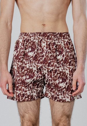 Mandlig overkrop iført løse shorts med elastisk linning i brun-hvidt abstrakt plettet mønster, stående mod ensfarvet baggrund.