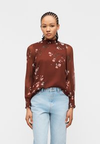 Femme aux cheveux tressés portant un chemisier marron à fleurs transparent à manches longues, rentré dans un jean taille haute bleu clair, debout devant un fond blanc.