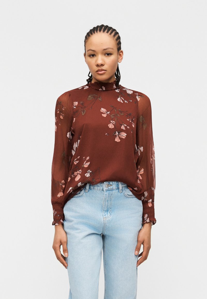 Femme aux cheveux tressés portant un chemisier marron à fleurs transparent à manches longues, rentré dans un jean taille haute bleu clair, debout devant un fond blanc.