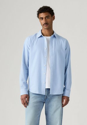 Mand med kort krøllet hår og overskæg iført en lyseblå skjorte med knapper over en hvid t-shirt og lyseblå jeans, stående foran en neutral baggrund.