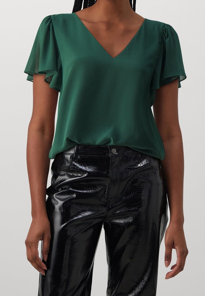 Personne portant un chemisier vert foncé à col en V avec des manches volantées et un pantalon noir brillant taille haute avec un motif texturé.