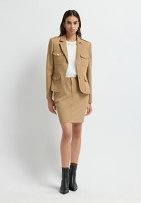 Mujer vestida con un blazer beige con cuatro bolsillos de solapa, falda lápiz a juego, blusa blanca, pendientes de aro y botas negras hasta el tobillo, de pie sobre un fondo blanco.