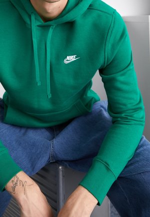 Osoba ubrana w zieloną bluzę Nike z kapturem i niebieskie dżinsy, siedząca z rękami spoczywającymi na kolanach, pokazująca cienki tatuaż na lewym nadgarstku.