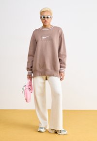 Túlméretes barna pulóver logóval, fehér csíkkal a mandzsettán, krém színű, bő szárú nadrág, szürke és rózsaszín sportcipő, valamint egy rózsaszín táska.