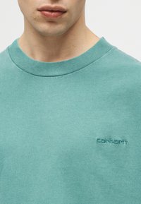 Κοντινό πλάνο ενός ατόμου που φοράει μπλούζα Carhartt σε τυρκουάζ απόχρωση με στρογγυλή λαιμόκοψη, εμφανίζοντας τα χείλη, τον λαιμό και το κεντημένο λογότυπο της μάρκας στο στήθος.