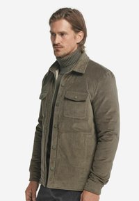 Mężczyzna w oliwkowej plecionce noszący kurtkę na matchingowy sweter z golfem, stojący na tle czystego białego tła.