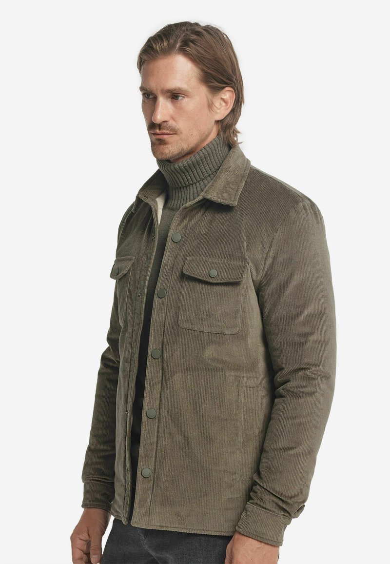 Mężczyzna w oliwkowej plecionce noszący kurtkę na matchingowy sweter z golfem, stojący na tle czystego białego tła.