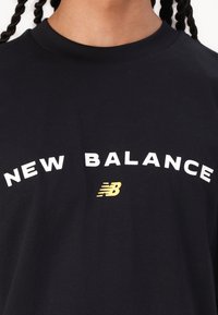 Melnā kokvilnas T-kreklā ir uzraksts "NEW BALANCE" treknrakstā baltās burtos un neliels dzeltenais logo, ar apaļu apkakli un īsām piedurknēm.