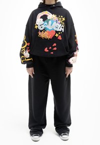 Sudadera corta negra con un diseño gráfico colorido que presenta corazones, nubes y estrellas. Combinada con pantalones negros holgados y zapatillas casuales.
