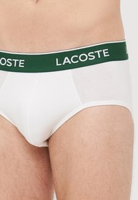 Weiße Baumwollslips mit einem grünen elastischen Bund, auf dem "LACOSTE" in weißer Schrift steht. Sanfte Textur, klassisches Slip-Design, enganliegende Form.