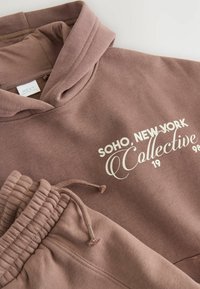 Felpa marrone con cappuccio e pantaloni jogger abbinati in morbido tessuto. Presenta un cappuccio con coulisse e una scritta bianca: "SOHO, NEW YORK Collective 1998."