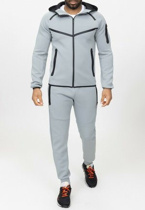 Ensemble de sport gris clair avec des accents noirs, poches zippées et capuche ; tissu texturé avec une coupe ajustée.
