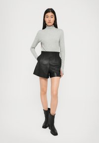 Donna in piedi con le mani in tasca, indossa una maglietta grigia a maniche lunghe, pantaloni shorts in pelle neri a vita alta e stivali neri alla caviglia.