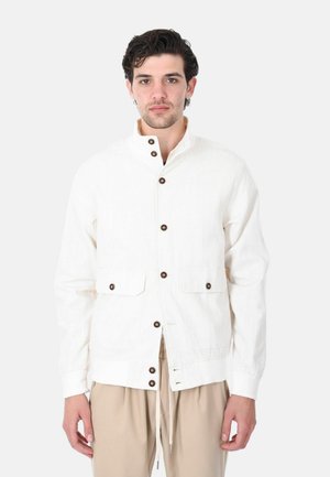 Uomo che indossa una giacca bianca abbottonata con tasche a pattina e pantaloni beige con coulisse, su uno sfondo chiaro uniforme.