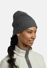 Gray knitted beanie with vertical ribbing, featuring a small logo patch. Worn with a light-colored fleece jacket and a high collar.