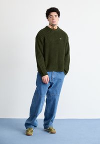 Σκούρο πράσινο πουλόβερ με υφή, στρογγυλή λαιμόκοψη, σε συνδυασμό με χαλαρές ανοιχτόμπλε τζιν παντελόνες και πράσινα αθλητικά παπούτσια με καφέ λεπτομέρειες.