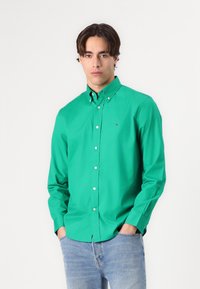 Camicia verde con bottoni e colletto, realizzata in tessuto morbido. Presenta un piccolo logo sul petto e maniche lunghe. Abbinata a jeans azzurri chiari.