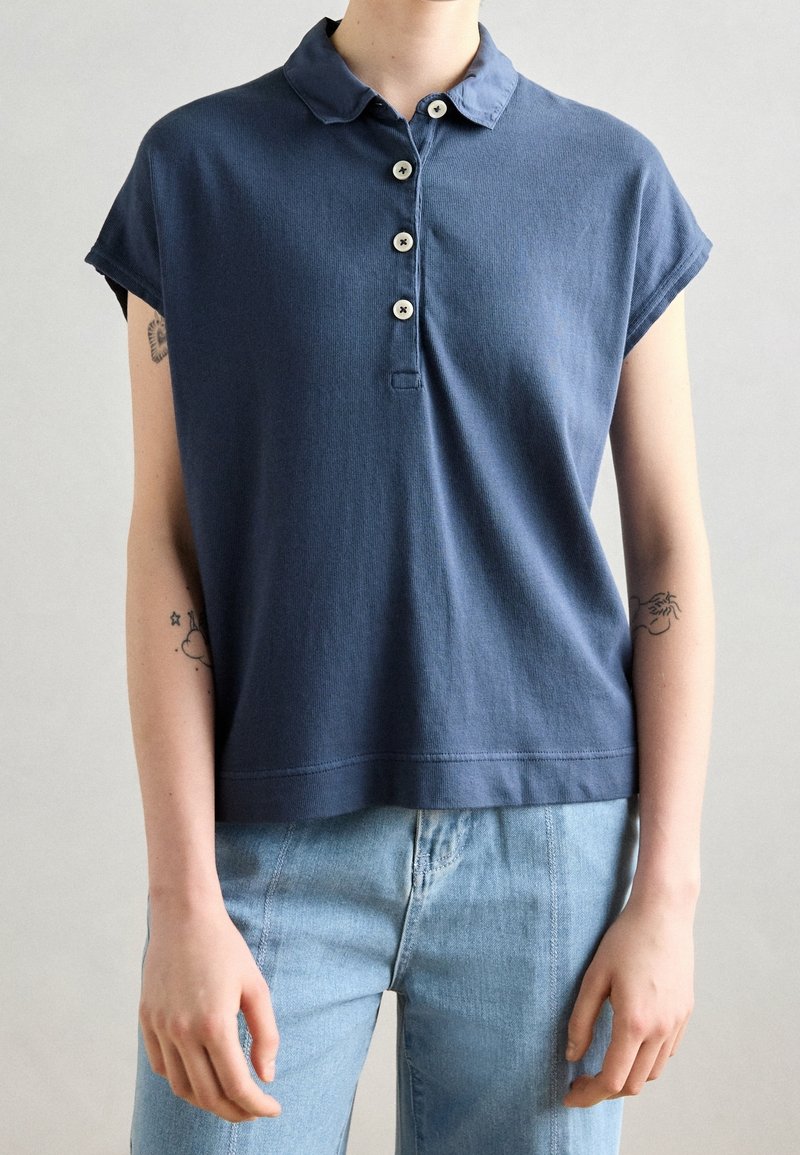 Personne portant une chemise bleu marine à manches courtes avec col et quatre boutons, et un jean bleu clair, debout devant un fond uni.