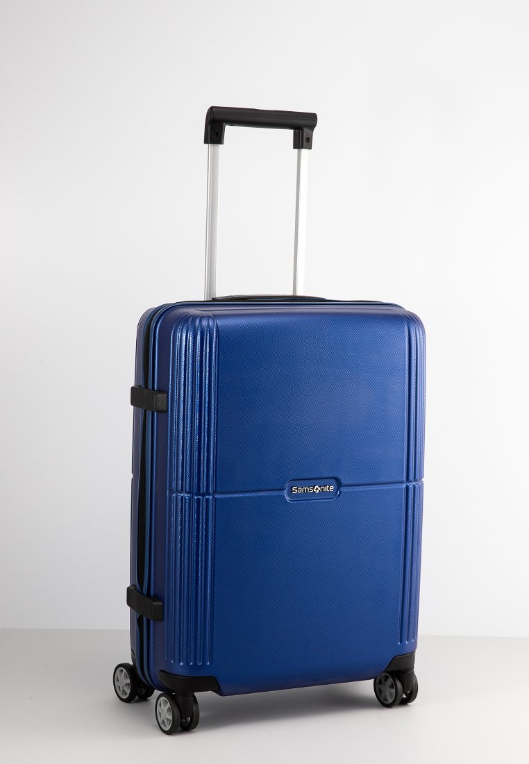 Maleta rígida azul con textura acanalada vertical, mango retráctil y cuatro ruedas negras. Presenta el logo de Samsonite en la parte frontal.