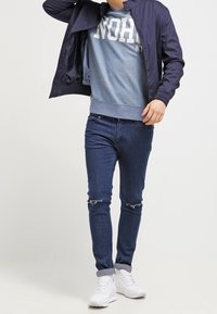 Navy bomber jacket over a blue graphic sweatshirt, paired with dark denim jeans featuring distressed knees and white sneakers.