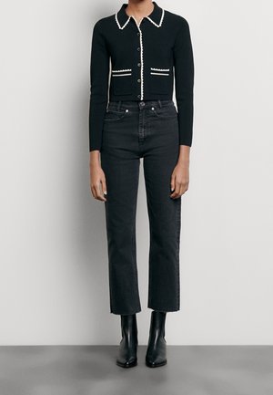 Zwarte kortgeknipte cardigan met witte biezen en zakken, gecombineerd met hoge zwarte jeans. Gekocht met spitse zwarte enkellaarsjes.