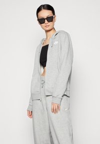Grå hettejakke med glidelås, hette og frontlommer, med en hvit Nike-logo. Kombinert med matchende grå joggebukser. Svart crop top under.